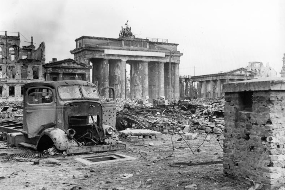 An old photograph of Brandenburg Gate; ruins of buildings and broken motor vehicles and several piles of debris made of bricks and mortar and metal in the background.